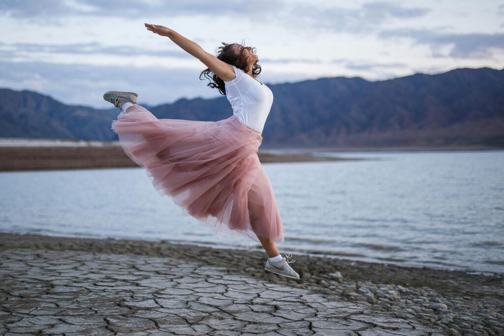 A woman joyfully jumps with outstretched arms by the lake with mountains in the background during the day.
